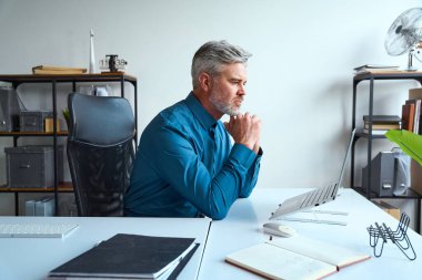 Modern ofis ortamında çalışan profesyonel bir adam, dizüstü bilgisayarla çalışmaya odaklanmış, kendini işine adamış, planlama ve verimliliği aktarıyor..