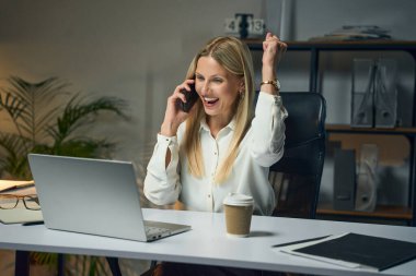 İş kadını telefonda konuşurken başarıyı kutluyor, dizüstü bilgisayar ve kahve kupası ile masada oturuyor, modern ofis çevreciliği, profesyonellik, verimlilik, kariyer hedefleri, motivasyon