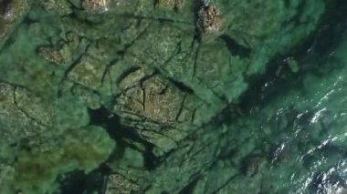 İnsansız hava aracıyla kayalık bir sahilde denizin şeffaf yeşil sularının üzerinde çekilmiş bir video.