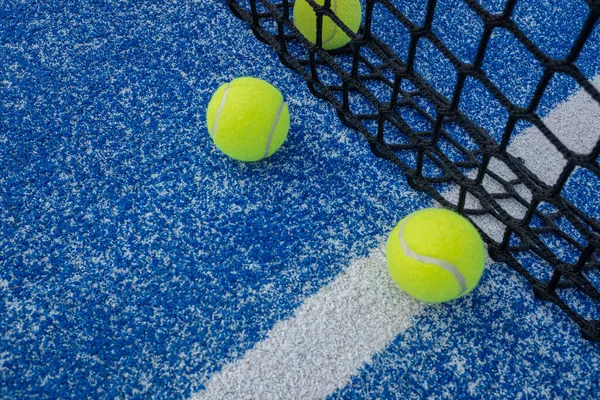 Three yellow balls on floor in front and behind of paddle net in blue court outdoors. Padel tennis