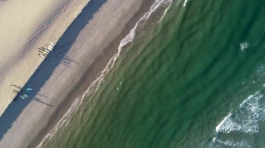 Bir grup sörfçünün sörf tahtaları ve yaz sporları konseptiyle sahil boyunca yürüdüğü drone hava görüntüsü.