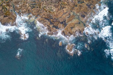 Turkuaz deniz yüzeyi, beyaz deniz köpüğü ve kıyıya vuran kayalar. İnsansız hava aracı ile üstten aşağı görünüm
