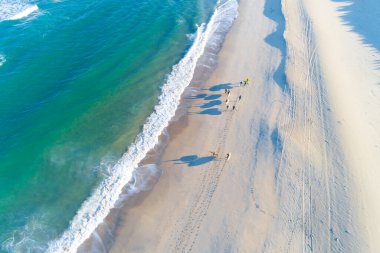 Bir grup sörfçü, sörf tahtaları ve insansız hava aracıyla kumsalda yürüyor..