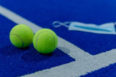 Mavi bir tenis kortunun yüzeyinde cerrahi maskenin yanında iki raket tenis topu. Salgın hastalıklarda spor.