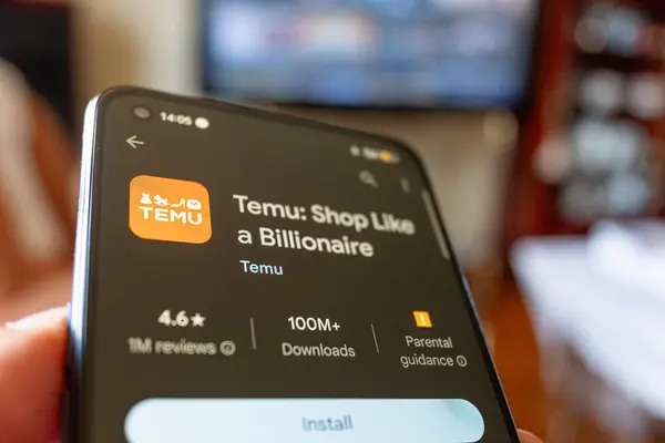 Madrid, İspanya 08-10-2024: Temu uygulama yükleyicisiyle akıllı telefon tutan kişinin eli, oturma odasında televizyonun önünde oturuyor.