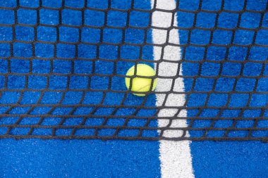 Parlak sarı tenis topu canlı bir mavi kortta ağa takıldı, top ve oyun yüzeyi arasındaki zıtlığı vurguluyor.