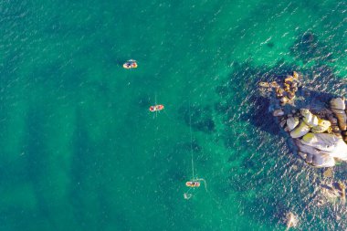 Kıyı sahili dron çekiminin hemen üstünde, şeffaf turkuaz suda yüzen tekneler.