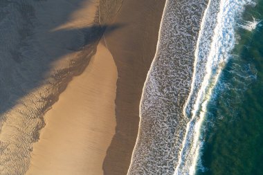 Tropikal bir sahil üzerindeki hava aracının yukarıdan görünüşü turkuaz su, altın kum, gün batımında köpüklü dalgalar.