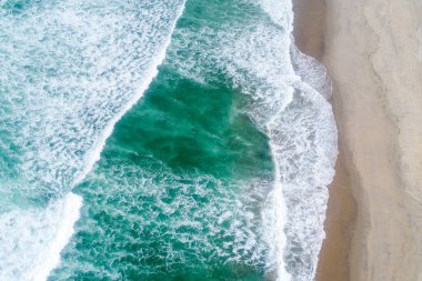 Geniş kıyı manzarası canlı yeşil dalgalar güneş ışığıyla aydınlanan sahile çarpıyor. Bir İHA 'nın havadan görüntüsü.