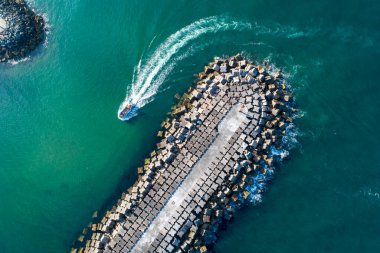 Turkuaz su yaz tatili konseptindeki büyük taştan bir dalgakıranın yanındaki sürat teknesinin hava görüntüsü.