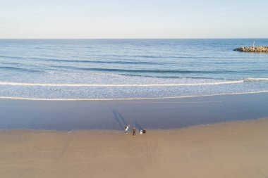 Gün batımında sörfçülerin olduğu geniş kumlu bir sahil manzarası..