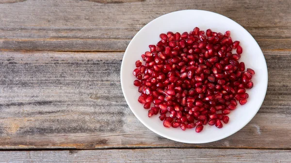 stock image Ripe pomegranate grains in a white  plate isolated on a wooden background. Top view, flat lay.