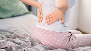 Genç bir kadın yatakta oturuyor ve sırt ağrısı çekiyor.