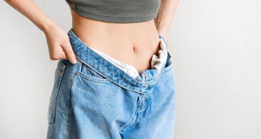 Diyet ve kilo verme konsepti. Büyük kotlu genç bir kadın açık gri arka planda ince vücudunu gösteriyor..