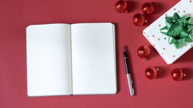 Kırmızı arka planda boş bir defter, hediyeler ve süslemeler bulunan Noel temalı bir masanın yukarıdan aşağı görünümü