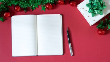 Kırmızı arka planda boş bir defter, hediyeler ve süslemeler bulunan Noel temalı bir masanın yukarıdan aşağı görünümü