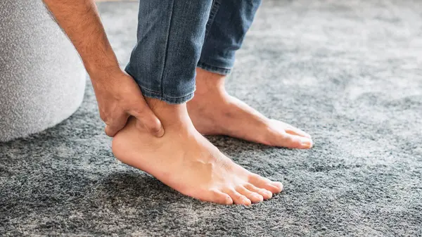 Ayak ağrısı konsepti, masaj yapan genç adamın eli, topuk bölgesinin masajı, kanepede acı çeken adam, evdeki kanepe..