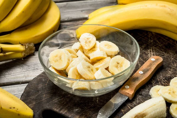 stock image Bananas and banana slices in a plate on a black chopping Board with a knife. On a wooden background.