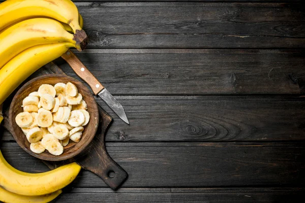 stock image Bananas and banana pieces in a wooden plate on a cutting Board with a knife. On a black wooden background.