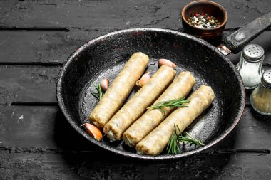 Biberiye ve sarımsak ile bir tavada çiğ sosis. Rustik siyah arka plan üzerine.