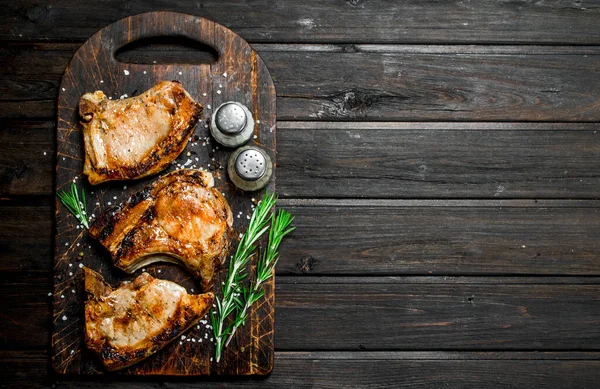 stock image Grilled pork steaks with rosemary and salt on an old board. On a wooden background.