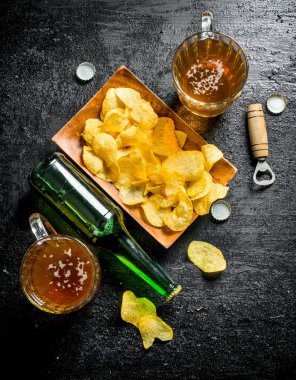 Bardaklarda bira ve tabakta bir şişe cips. Siyah rustik arka plan üzerinde