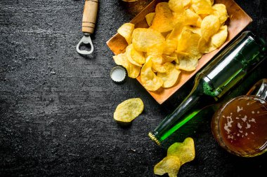 Bardaklarda bira ve tabakta bir şişe cips. Siyah rustik arka plan üzerinde