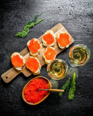 Kesme tahtasında kırmızı havyar ve bardakta beyaz şarap olan sandviçler. Siyah rustik arka plan üzerinde