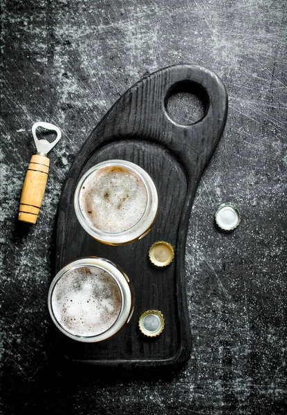 Stock image Beer in glasses on a black cutting Board with opener and covers. On black rustic background