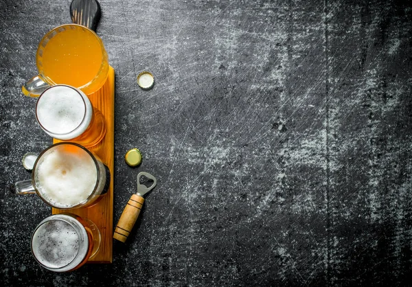 stock image Different glasses of beer on a wooden cutting Board with opener. On black rustic background