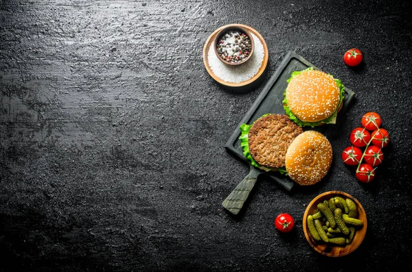 stock image Burgers on a cutting Board with gherkins and spices in bowls. On rustic background