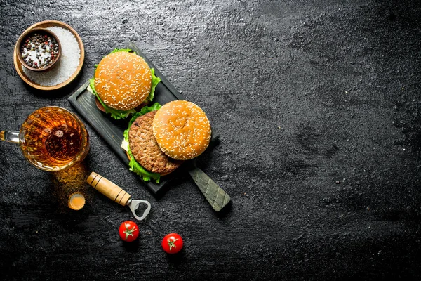 stock image Burgers with beer in a glass and tomatoes. On black rustic background