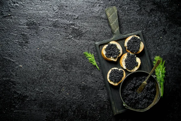 stock image Black caviar in a bowl and sandwiches with caviar on a cutting Board with dill. On black rustic background