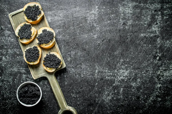 stock image Sandwiches with black caviar on a cutting Board and caviar in a bowl. On black rustic background