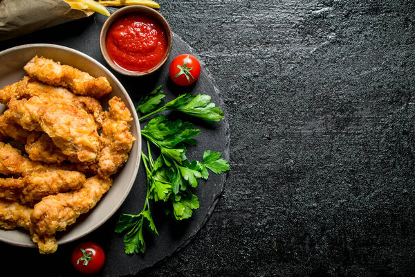 Chicken strips with French fries, greens and tomato sauce. On black rustic background
