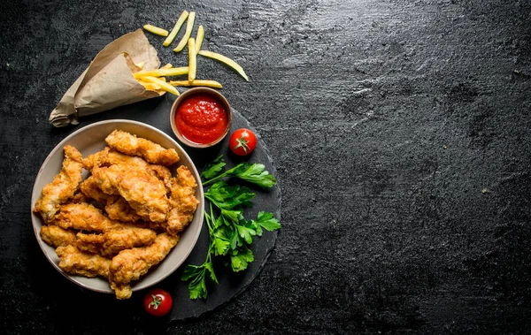 stock image Chicken strips in a bowl with parsley, tomatoes, sauce and French fries. On black rustic background