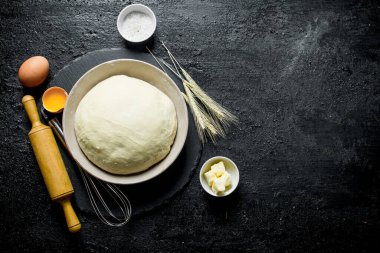 Bir oklava, sivri ve yumurta ile bir kapta hamur. Siyah rustik arka plan üzerinde