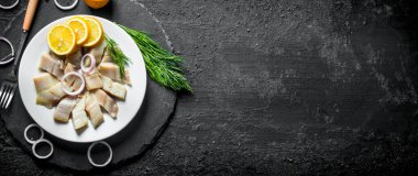 Dereotu, limon ve soğan halkaları ile dilimlenmiş tuzlu ringa balığı. Siyah rustik arka plan üzerinde