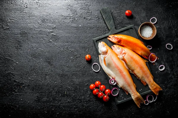 stock image Raw fish trout on a cutting Board with onion rings and cherry tomatoes. On black rustic background