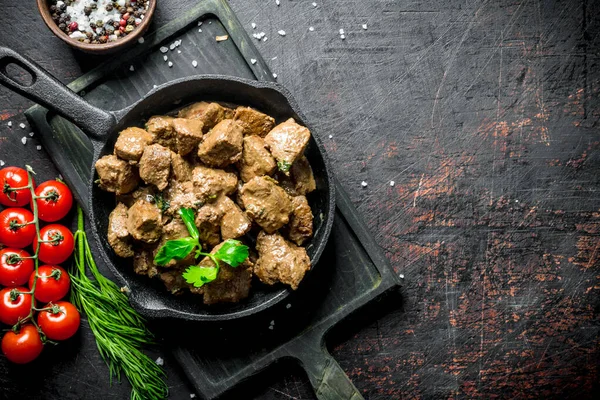 stock image Pieces of cooked liver in a frying pan on a cutting Board with tomatoes, herbs and spices. On dark rustic background