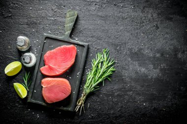 Biberiye, baharat ve limon dilimleri ile bir kesme Kurulu taze çiğ ton balığı biftek. Siyah rustik arka plan üzerinde