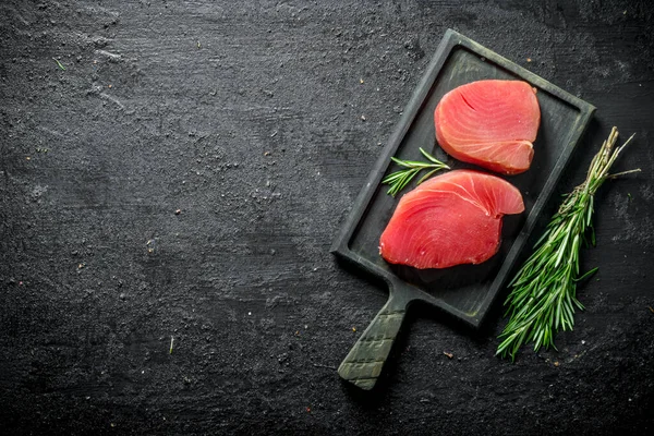 stock image Pieces of raw tuna on a cutting Board with a bunch of rosemary. On black rustic background