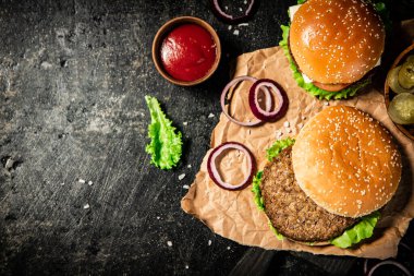 Burger on a cutting board with onion rings, tomato sauce and lettuce. On a black background. 