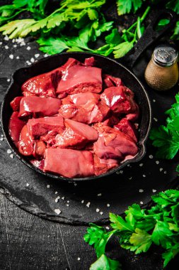 Pieces of raw liver in a frying pan with parsley and spices. On a black background. High quality photo