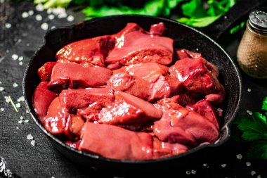 Pieces of raw liver in a frying pan with parsley and spices. On a black background. High quality photo