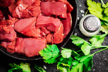 Pieces of raw liver in a frying pan with parsley and spices. On a black background. High quality photo
