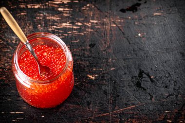 Red caviar in an open glass jar with a spoon. On a rustic dark background. High quality photo