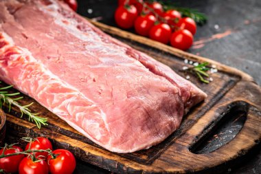 A piece of raw pork on a cutting board with tomatoes, spices and rosemary. On a rustic dark background. High quality photo