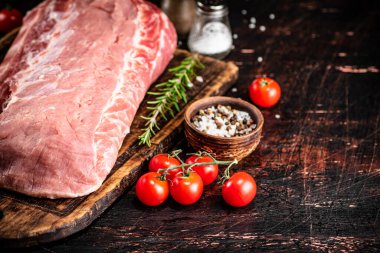 A piece of raw pork on a cutting board with tomatoes, spices and rosemary. On a rustic dark background. High quality photo