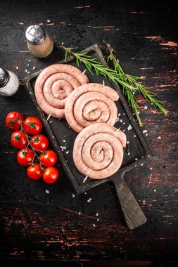 Raw sausages on a cutting board with a sprig of rosemary and tomatoes. On a rustic dark background. High quality photo